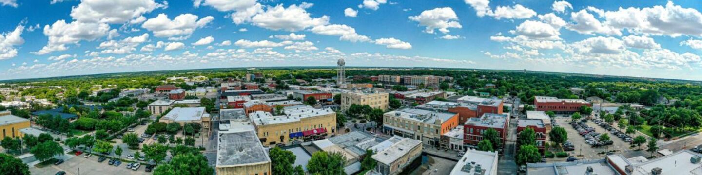 Aerial panoramic view of historic Downtown McKinney, Texas, featuring storefronts, tree-lined streets, the downtown water tower, and a blue sky with white clouds.
