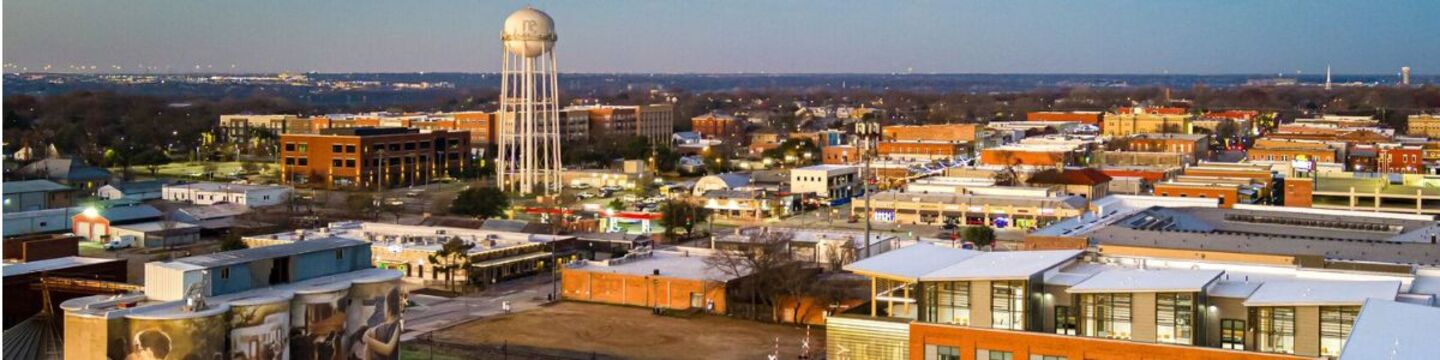 Aerial panoramic view of historic Downtown McKinney, Texas, featuring storefronts, tree-lined streets, the downtown water tower, and a blue sky with white clouds.