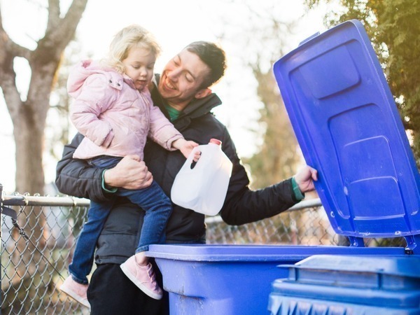 man holding child who is putting a milk carton in a recycling cart